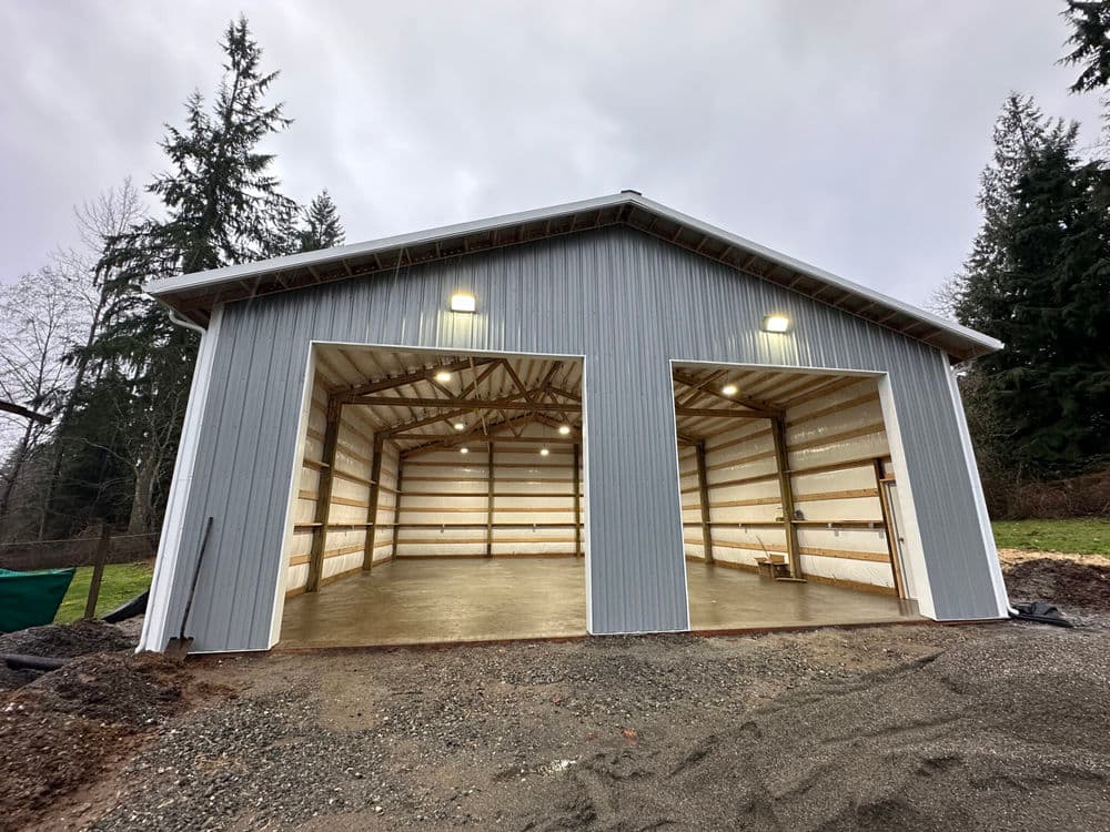 Modern metal storage barn with open garage doors, surrounded by trees and gravel.