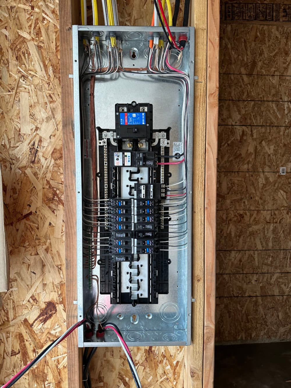 Electrical panel with circuit breakers and wiring, mounted on an unfinished wall.
