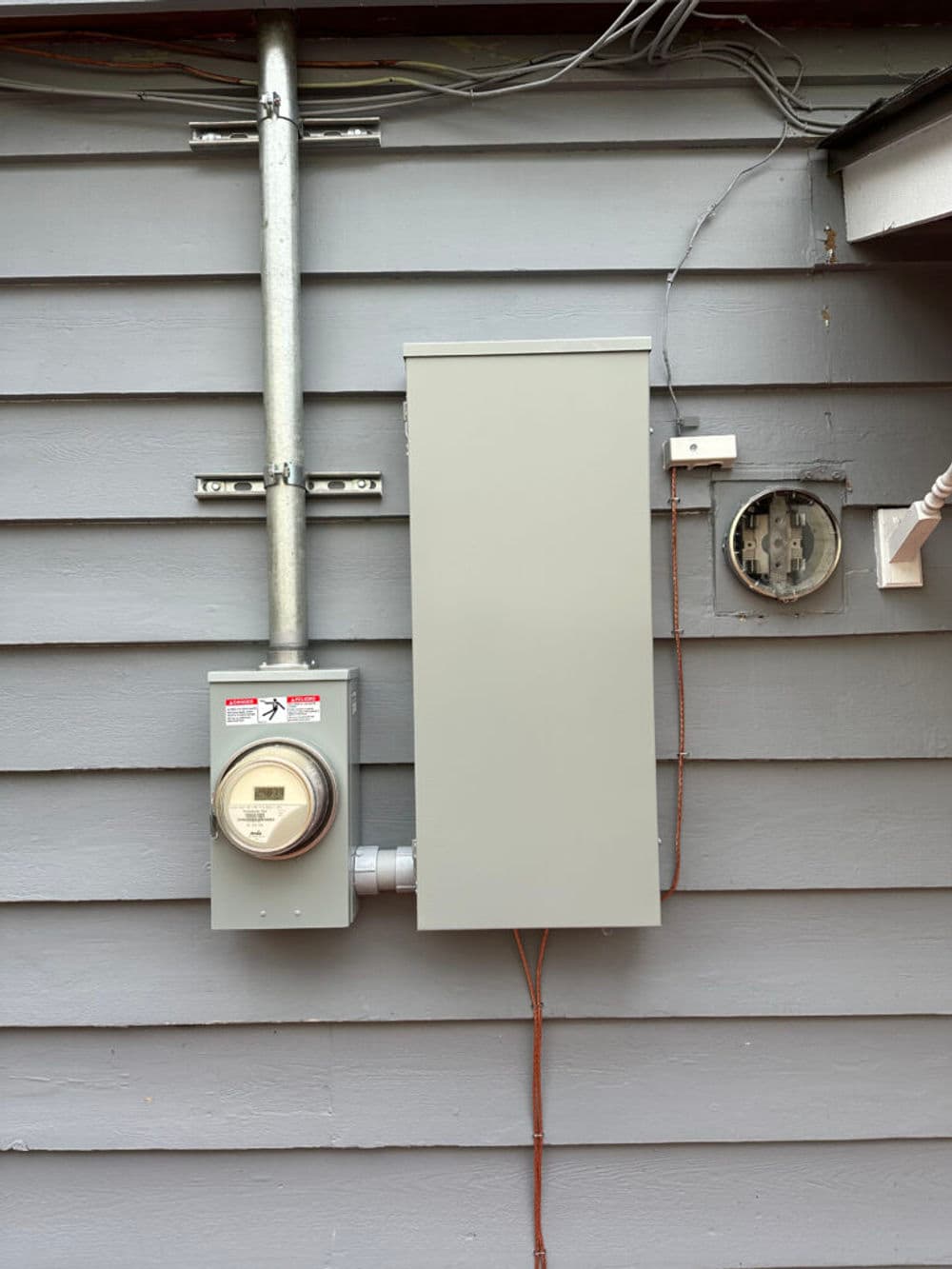 Electric meter and service panel mounted on gray wooden wall with exposed wiring.