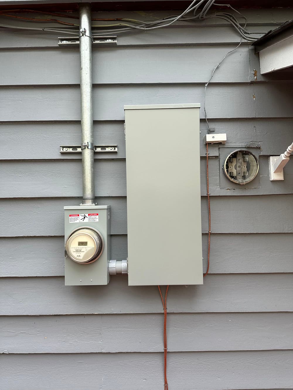 Electric meter and service panel on gray house exterior wall.