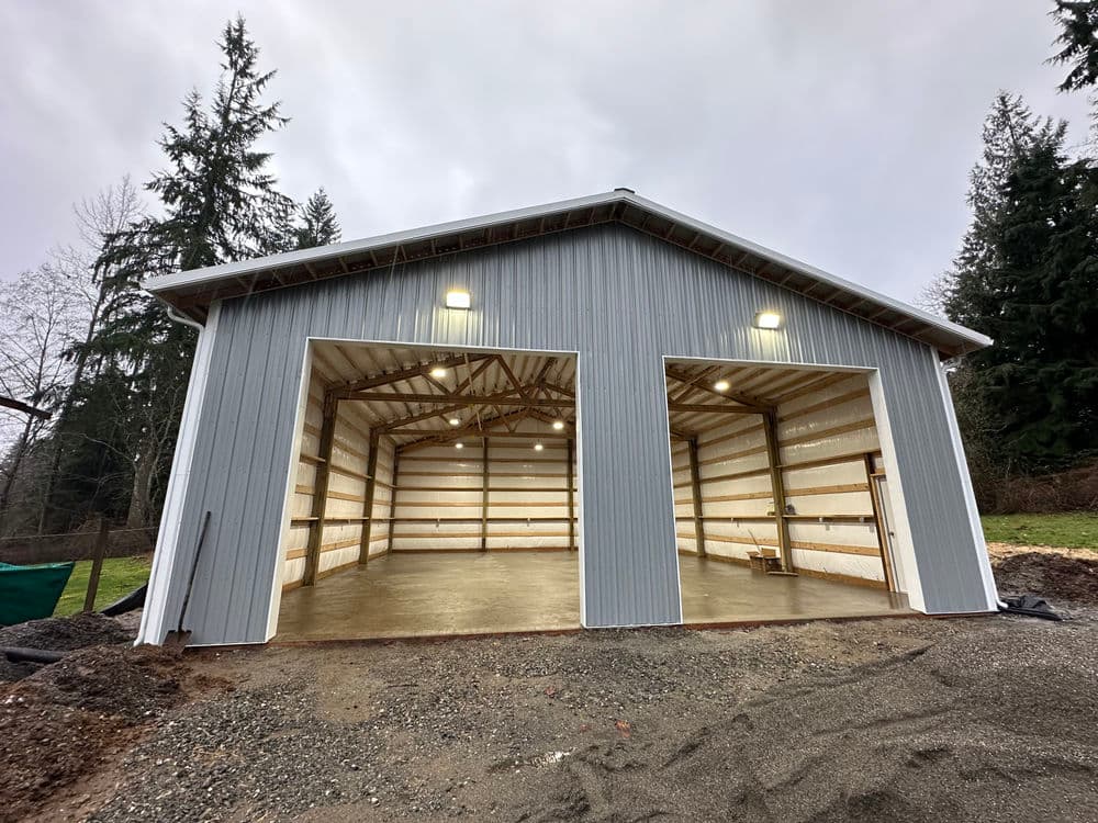 Modern metal barn with open entrances, LED lights, and gravel foreground, surrounded by trees.