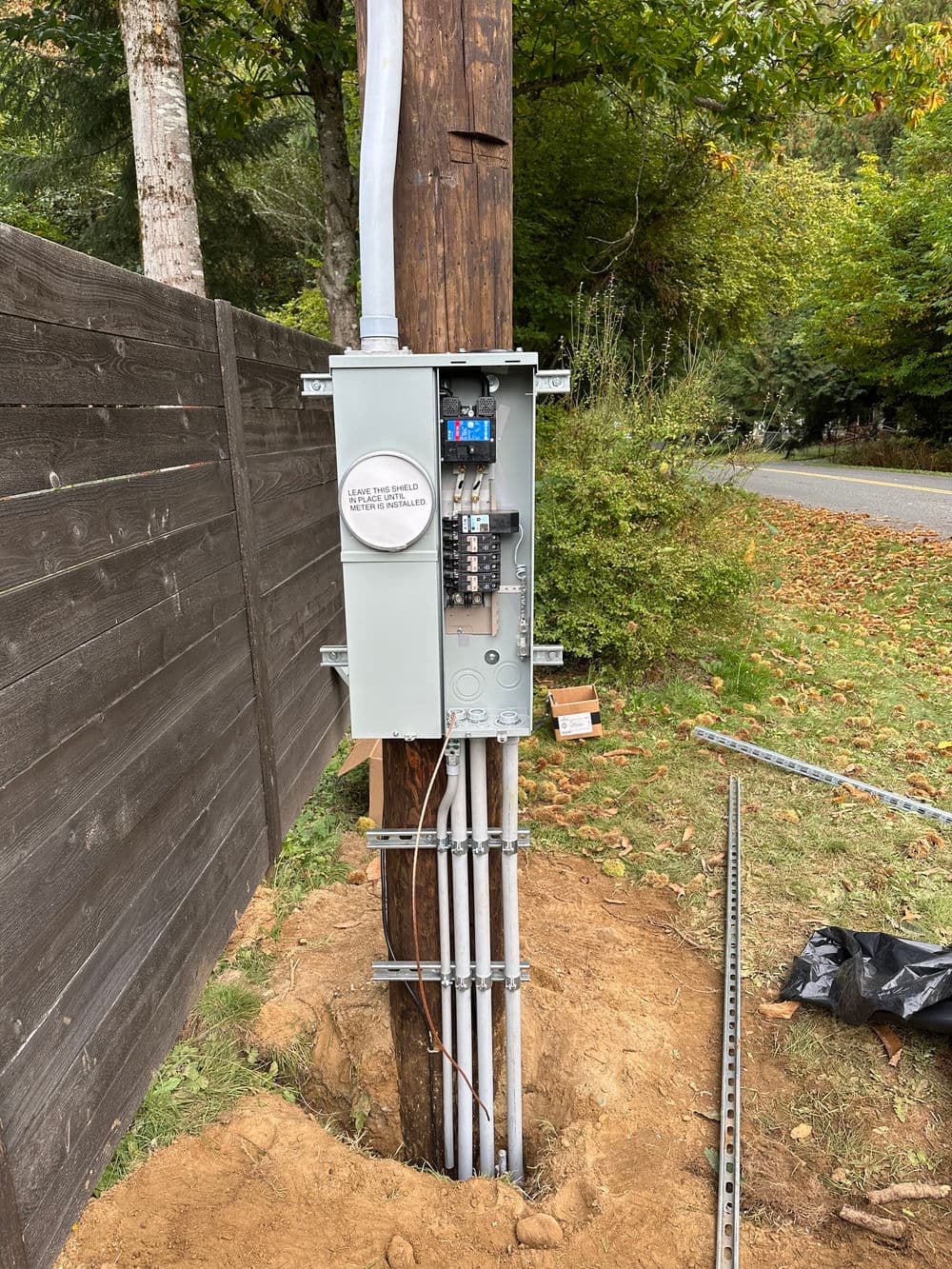 Utility pole with meter and electrical panel, surrounded by trees and dirt.